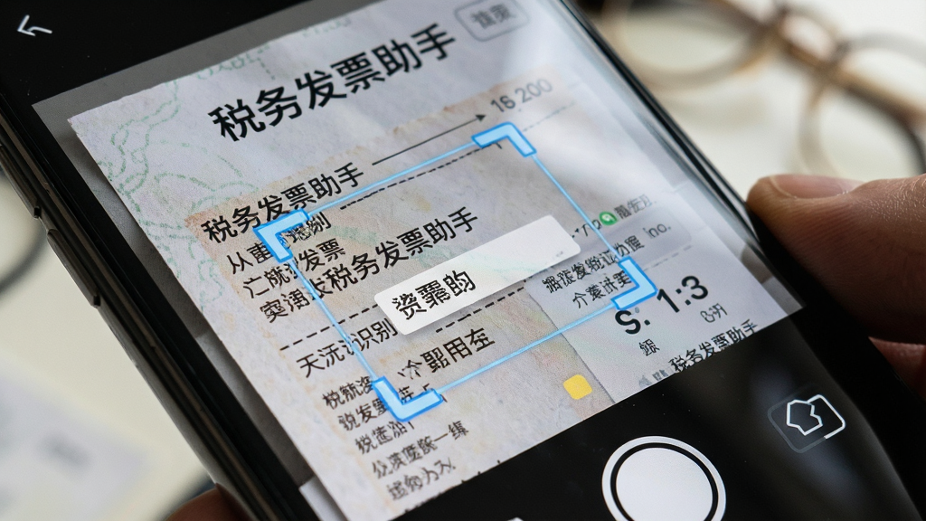 一张展示“税务发票助手”智能识别功能的细节图,主体是手机摄像头对准一张纸质发票,屏幕上显示识别框和正在提取的信息,布局为近景特写,排版突出识别过程,风格为真实场景与UI结合,细节上能看到发票纹理和屏幕反光,画质清晰,无多余元素,图片中有“税务发票助手”文字标识。