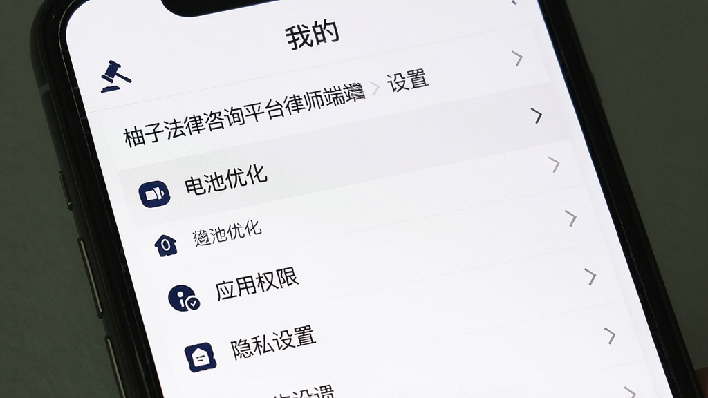 手机截图展示“柚子法律咨询平台律师端”的设置页面,页面中高亮显示“电池优化”选项和“应用权限”入口,屏幕上方有“我的”标题栏,下方列表包含通知管理、隐私设置等条目,界面风格简洁,以白色背景和深蓝色图标为主,画质清晰干净,无多余元素