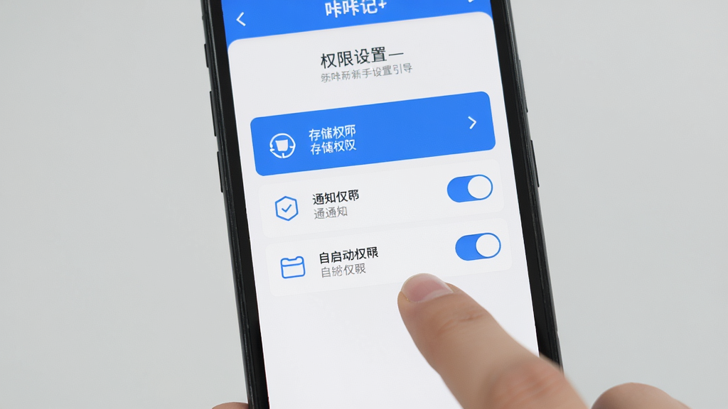 咔咔记新手设置引导界面展示,手机屏幕中显示咔咔记应用的权限设置页面,包含存储权限、通知权限、自启动权限的开关选项,界面风格简洁,蓝白配色,图标清晰,文字标签明确,整体布局清爽,画质干净,无多余元素,背景为浅灰色,画面中有一只手正在点击屏幕