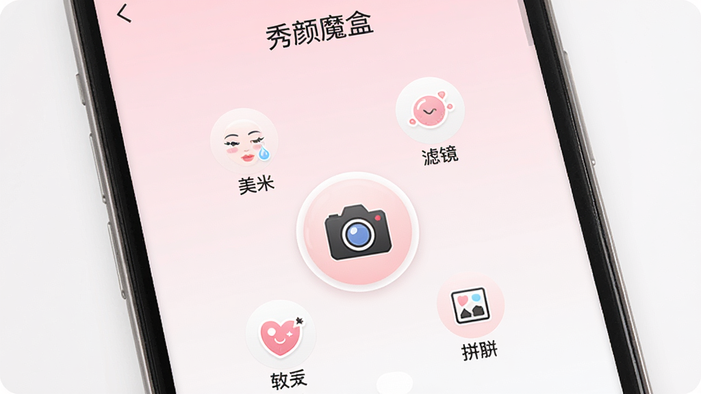 一张展示智能手机屏幕的图片,屏幕中显示着“秀颜魔盒”APP的主界面,界面以柔和的粉白色调为主,中心位置有一个圆形的相机图标,周围环绕着美颜、滤镜、贴纸、拼图四个功能按钮,背景是干净的浅色渐变,整体风格清新简约,画质干净清晰,无多余元素