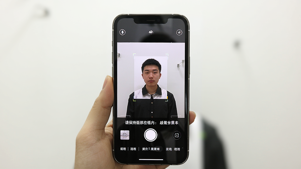 一张手机屏幕显示证件照拍摄院的拍摄界面,画面中有一个人站在白墙前,手机屏幕上显示取景框和“请保持面部在框内”的提示文字,下方有拍摄按钮和规格选择栏,整体光线明亮,风格简洁实用,画质清晰,无多余元素