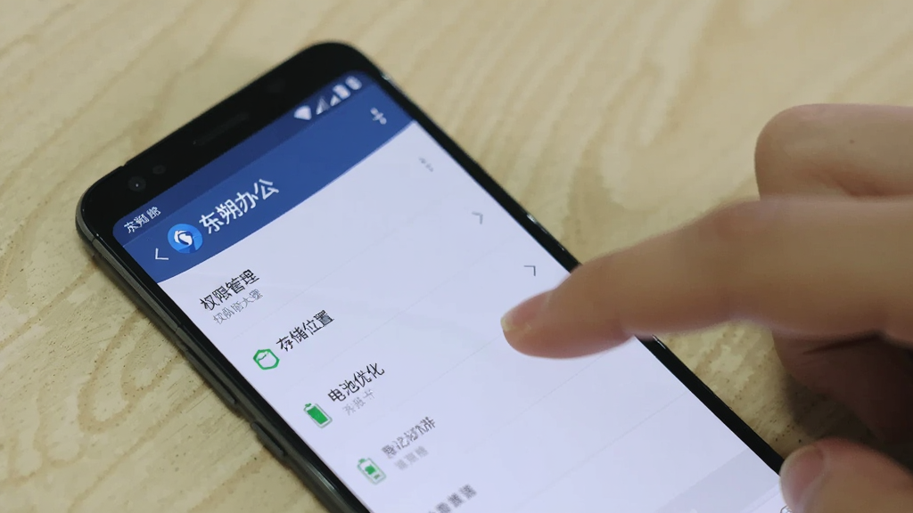 一部安卓手机显示“东朔办公”的设置页面,页面中有“权限管理”“存储位置”“电池优化”等选项,手机旁边有一只手正在点击屏幕,背景是浅色桌面,画质清晰,风格写实